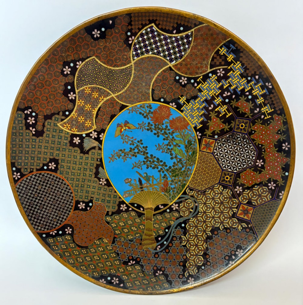 A circular Japanese Meiji period cloisonne charger featuring a sky-blue central medallion with chrysanthemums, a butterfly, and a frog, framed by geometric enamel patterns.