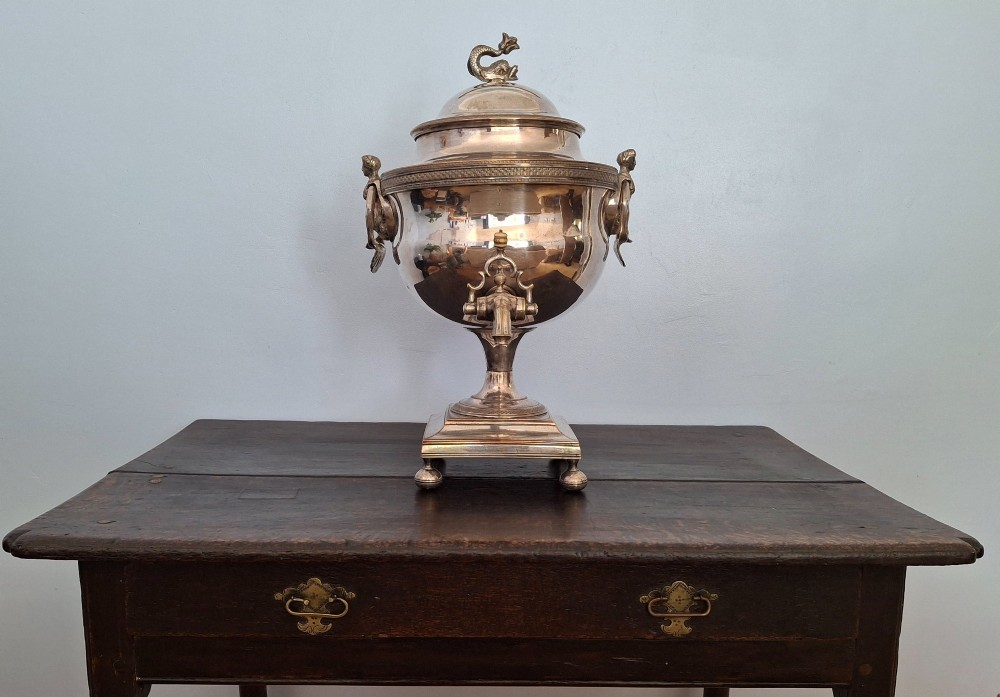 An antique silver-plated Regency tea urn featuring a top dolphin finial, classical figure side handles, a front spigot, and a square base supported by ball feet. View II