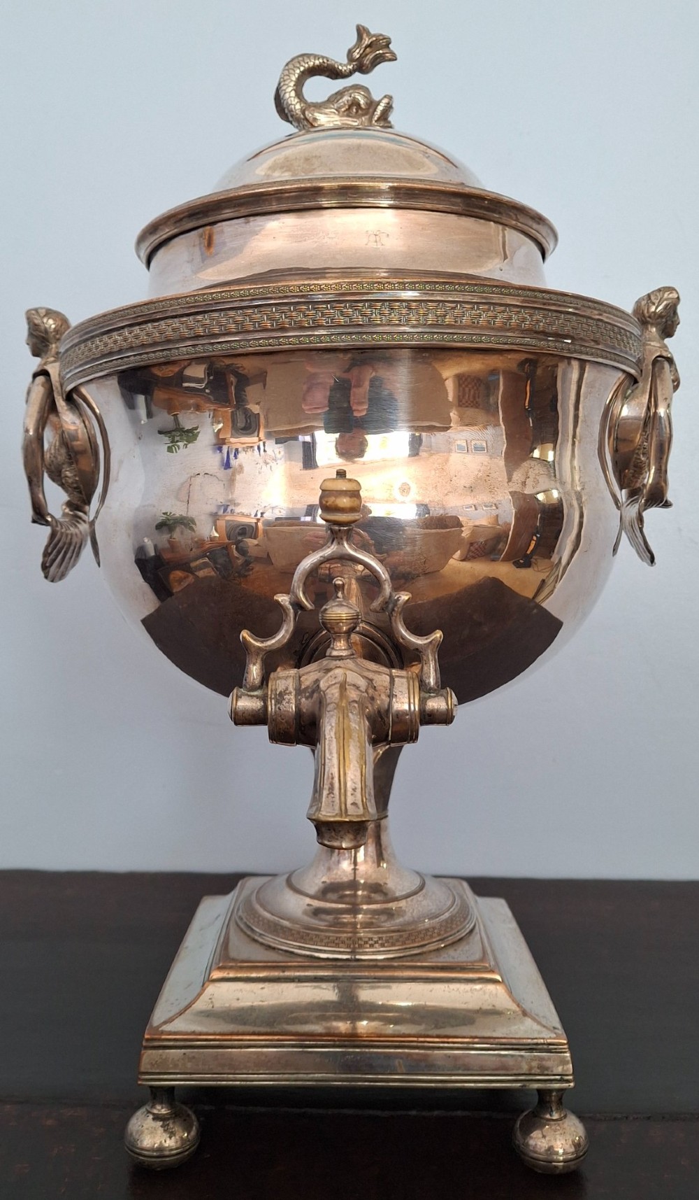 An antique silver-plated Regency tea urn featuring a top dolphin finial, classical figure side handles, a front spigot, and a square base supported by ball feet.