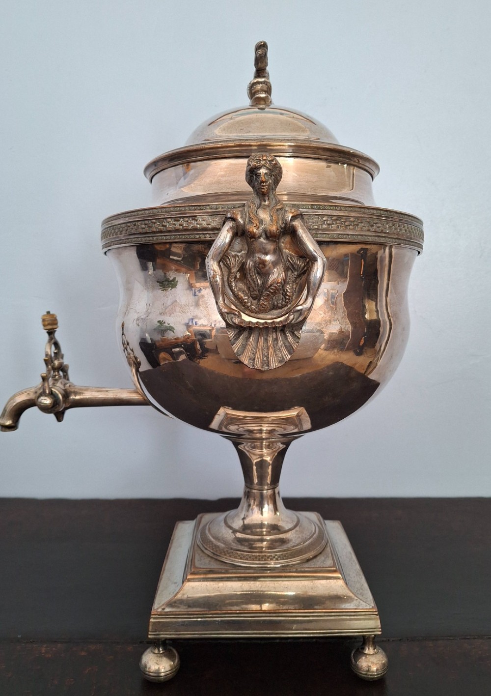 An antique silver-plated Regency tea urn featuring a top dolphin finial, classical figure side handles, a front spigot, and a square base supported by ball feet. View IV