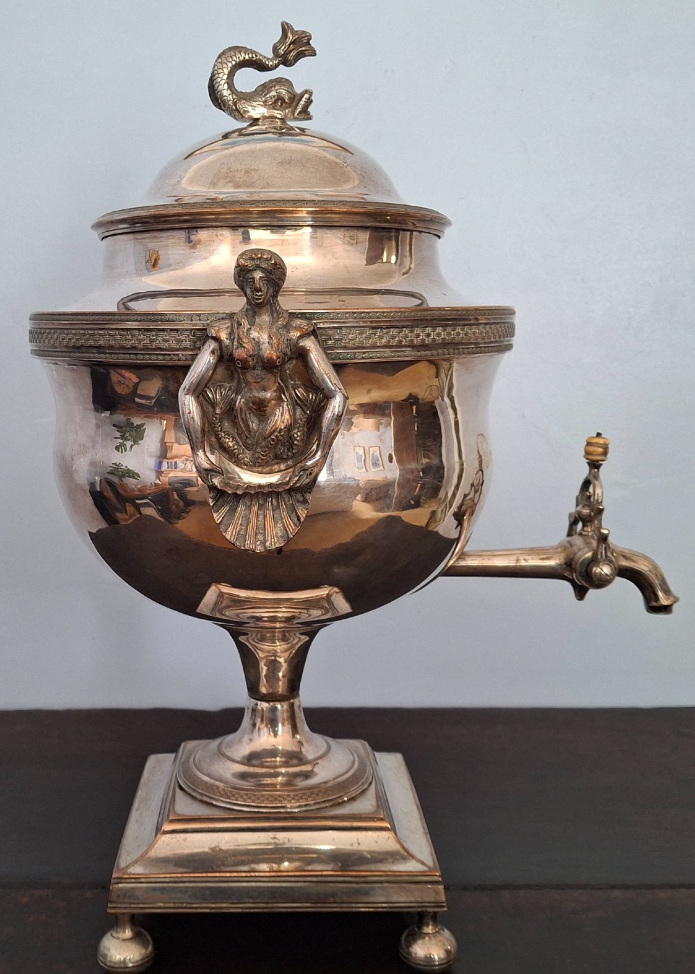 An antique silver-plated Regency tea urn featuring a top dolphin finial, classical figure side handles, a front spigot, and a square base supported by ball feet. View V