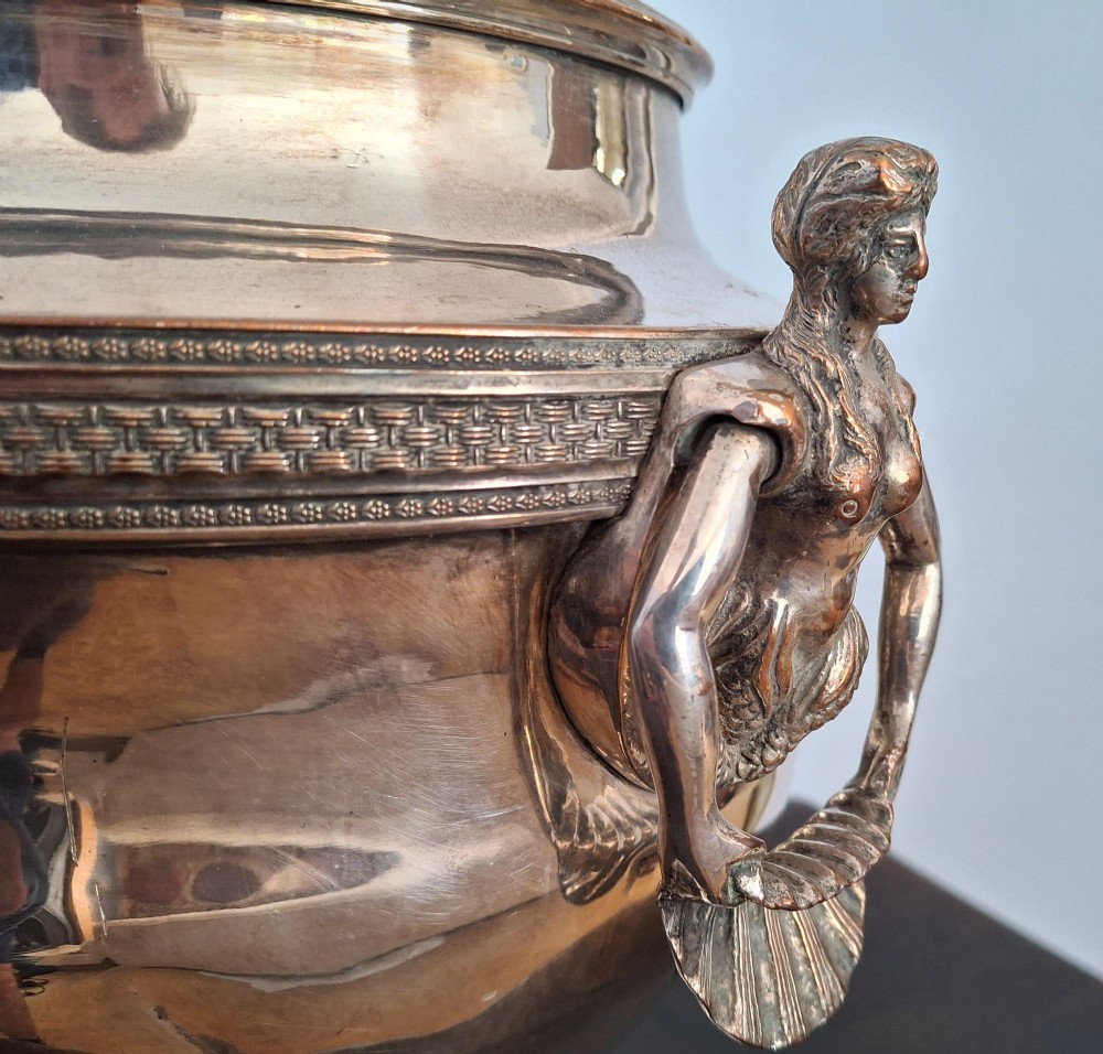 An antique silver-plated Regency tea urn featuring a top dolphin finial, classical figure side handles, a front spigot, and a square base supported by ball feet. View VI