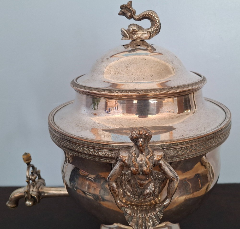 An antique silver-plated Regency tea urn featuring a top dolphin finial, classical figure side handles, a front spigot, and a square base supported by ball feet. View VII