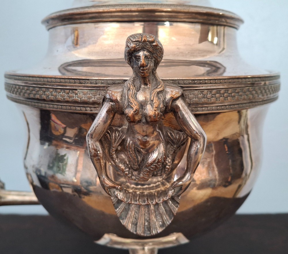 An antique silver-plated Regency tea urn featuring a top dolphin finial, classical figure side handles, a front spigot, and a square base supported by ball feet. View VIII