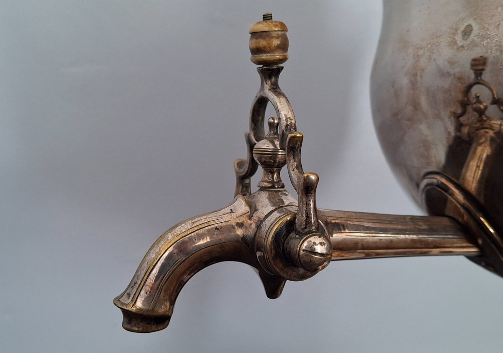 An antique silver-plated Regency tea urn featuring a top dolphin finial, classical figure side handles, a front spigot, and a square base supported by ball feet. View IX