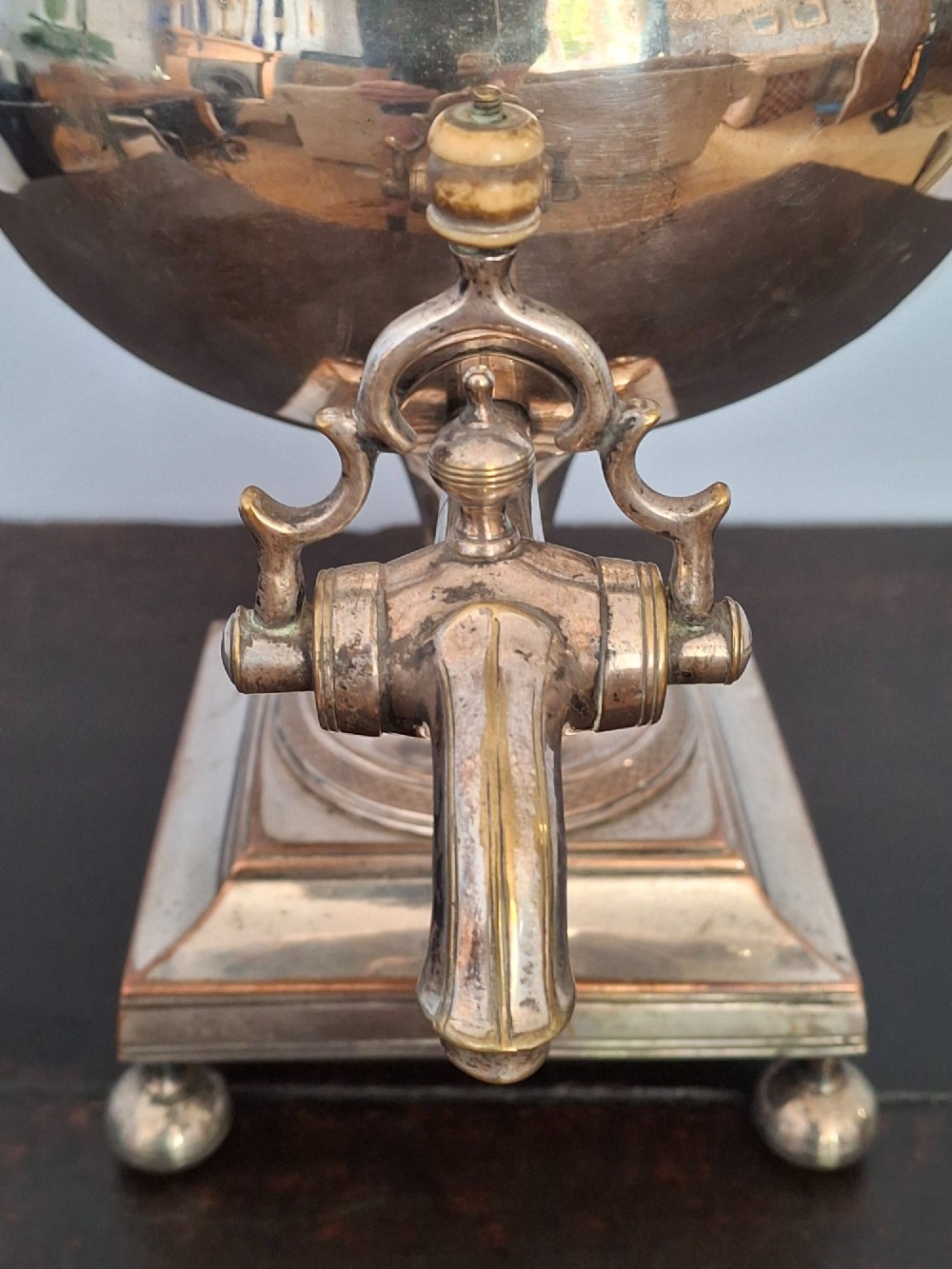 An antique silver-plated Regency tea urn featuring a top dolphin finial, classical figure side handles, a front spigot, and a square base supported by ball feet. View X