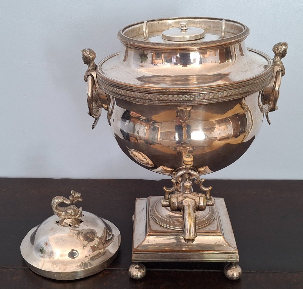 An antique silver-plated Regency tea urn featuring a top dolphin finial, classical figure side handles, a front spigot, and a square base supported by ball feet. View XI