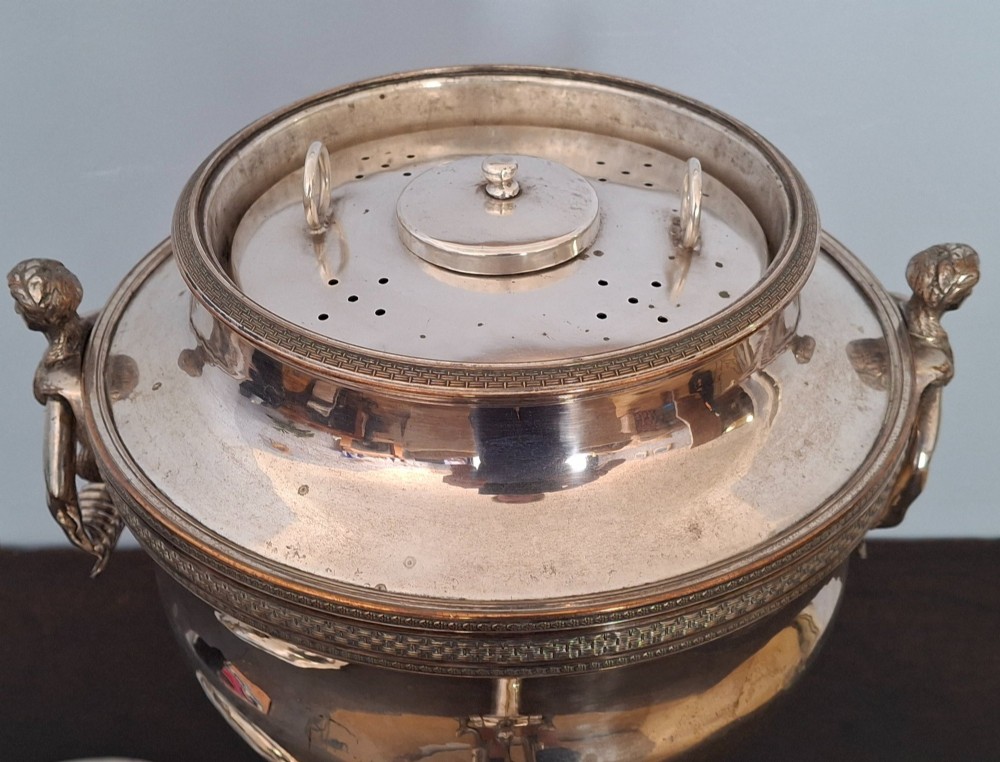 An antique silver-plated Regency tea urn featuring a top dolphin finial, classical figure side handles, a front spigot, and a square base supported by ball feet. View XII