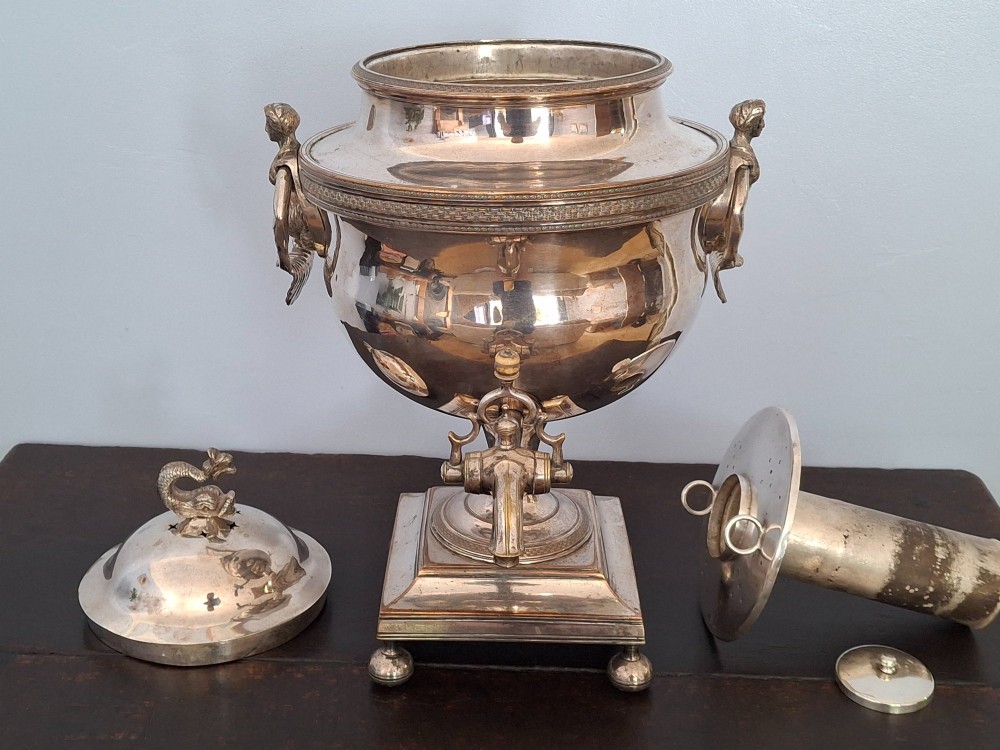 An antique silver-plated Regency tea urn featuring a top dolphin finial, classical figure side handles, a front spigot, and a square base supported by ball feet. View XIII