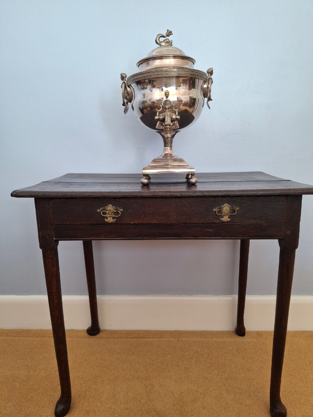 An antique silver-plated Regency tea urn featuring a top dolphin finial, classical figure side handles, a front spigot, and a square base supported by ball feet. View XV