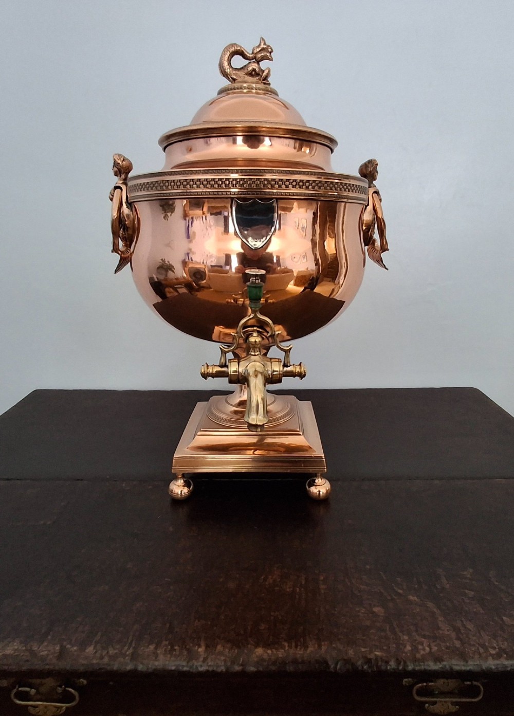 An antique polished copper and brass tea urn featuring a urn-shaped body, crest finial, side handles, and a spigot, resting on a square pedestal base.
