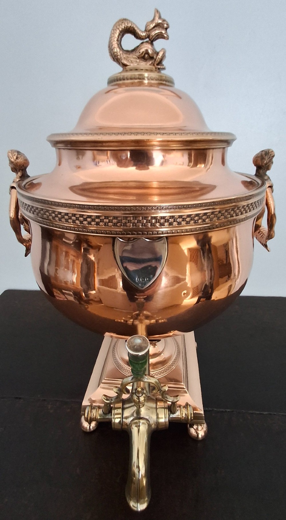 An antique polished copper and brass tea urn featuring a urn-shaped body, crest finial, side handles, and a spigot, resting on a square pedestal base. View II