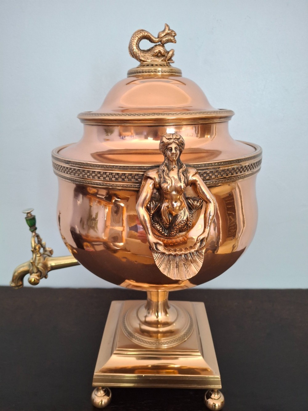 An antique polished copper and brass tea urn featuring a urn-shaped body, crest finial, side handles, and a spigot, resting on a square pedestal base. View IV