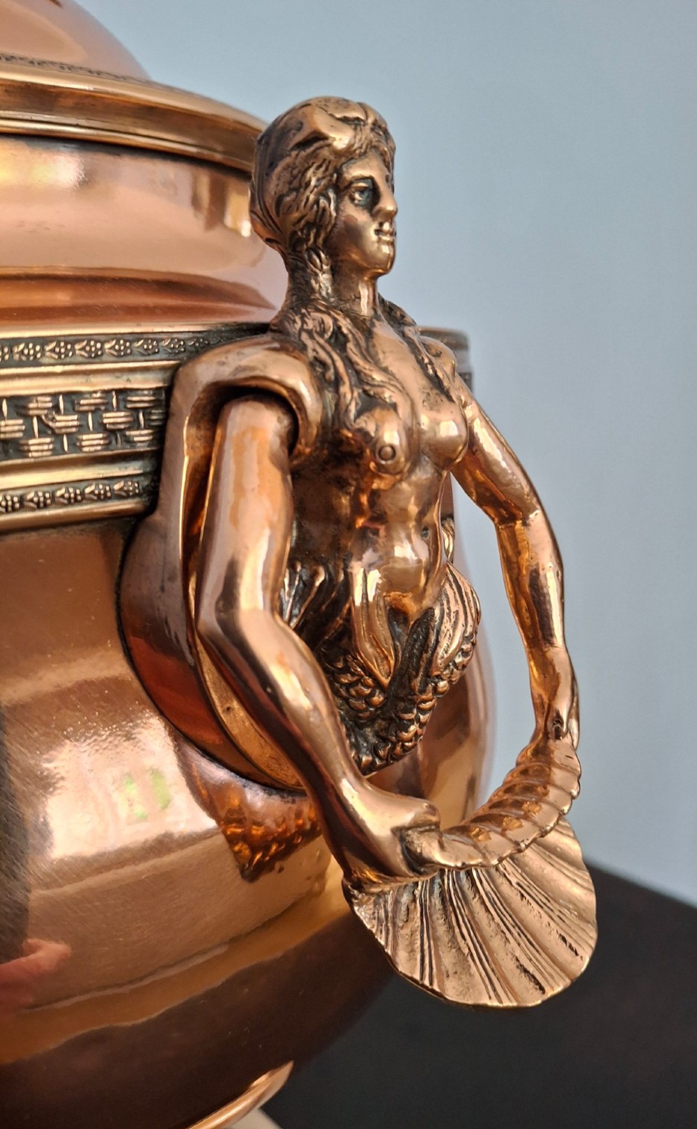 An antique polished copper and brass tea urn featuring a urn-shaped body, crest finial, side handles, and a spigot, resting on a square pedestal base. View VI