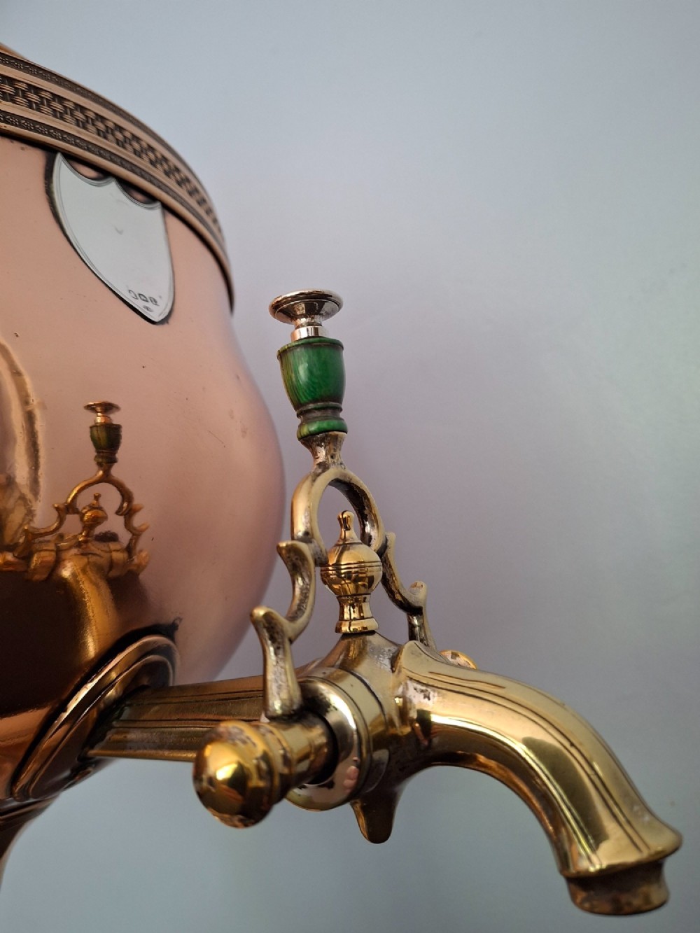 An antique polished copper and brass tea urn featuring a urn-shaped body, crest finial, side handles, and a spigot, resting on a square pedestal base. View VIII