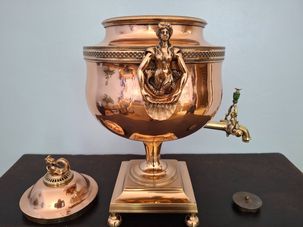 An antique polished copper and brass tea urn featuring a urn-shaped body, crest finial, side handles, and a spigot, resting on a square pedestal base. View IX