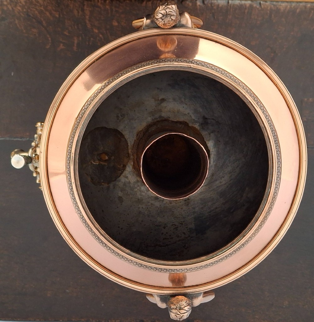 An antique polished copper and brass tea urn featuring a urn-shaped body, crest finial, side handles, and a spigot, resting on a square pedestal base. View X