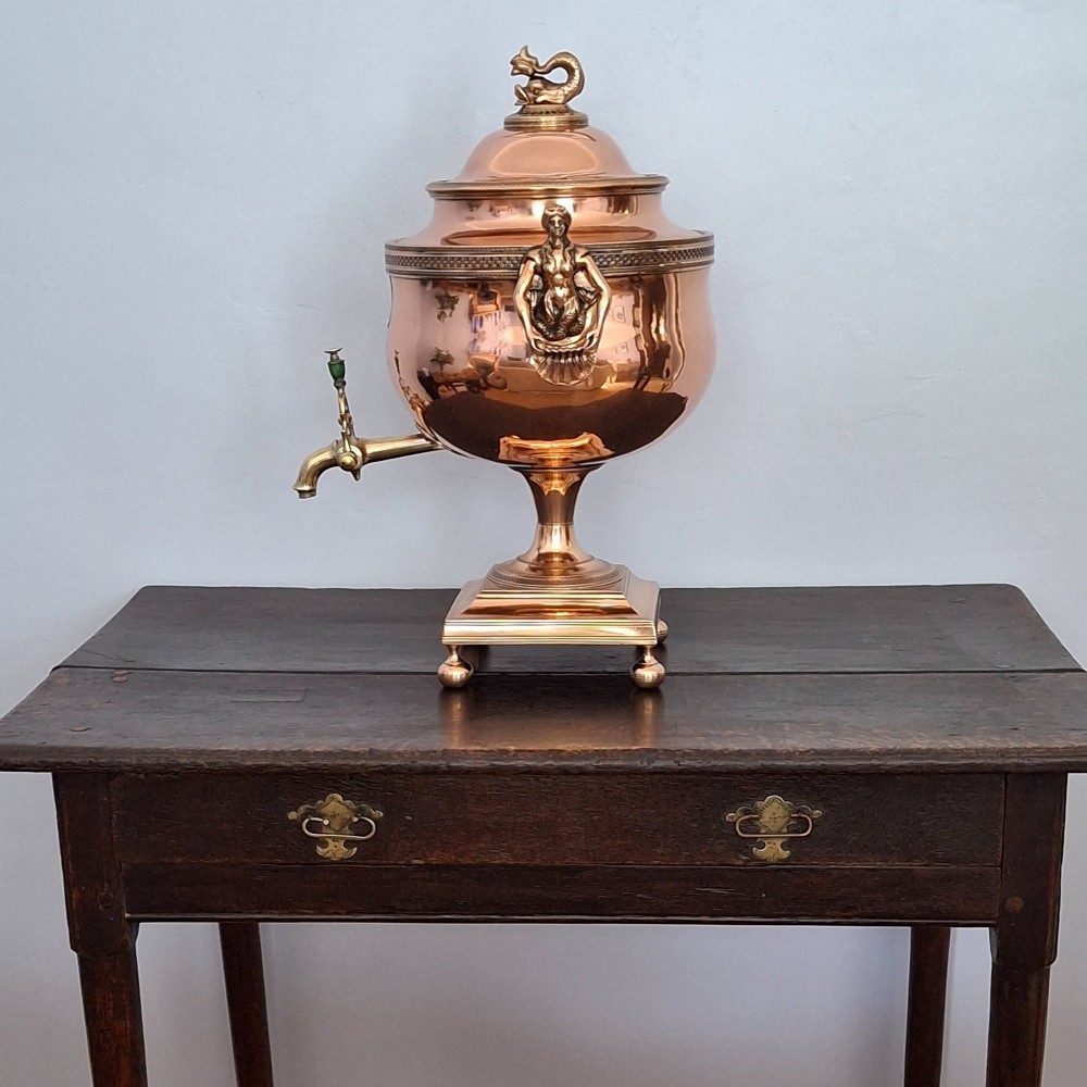 An antique polished copper and brass tea urn featuring a urn-shaped body, crest finial, side handles, and a spigot, resting on a square pedestal base. View XIV