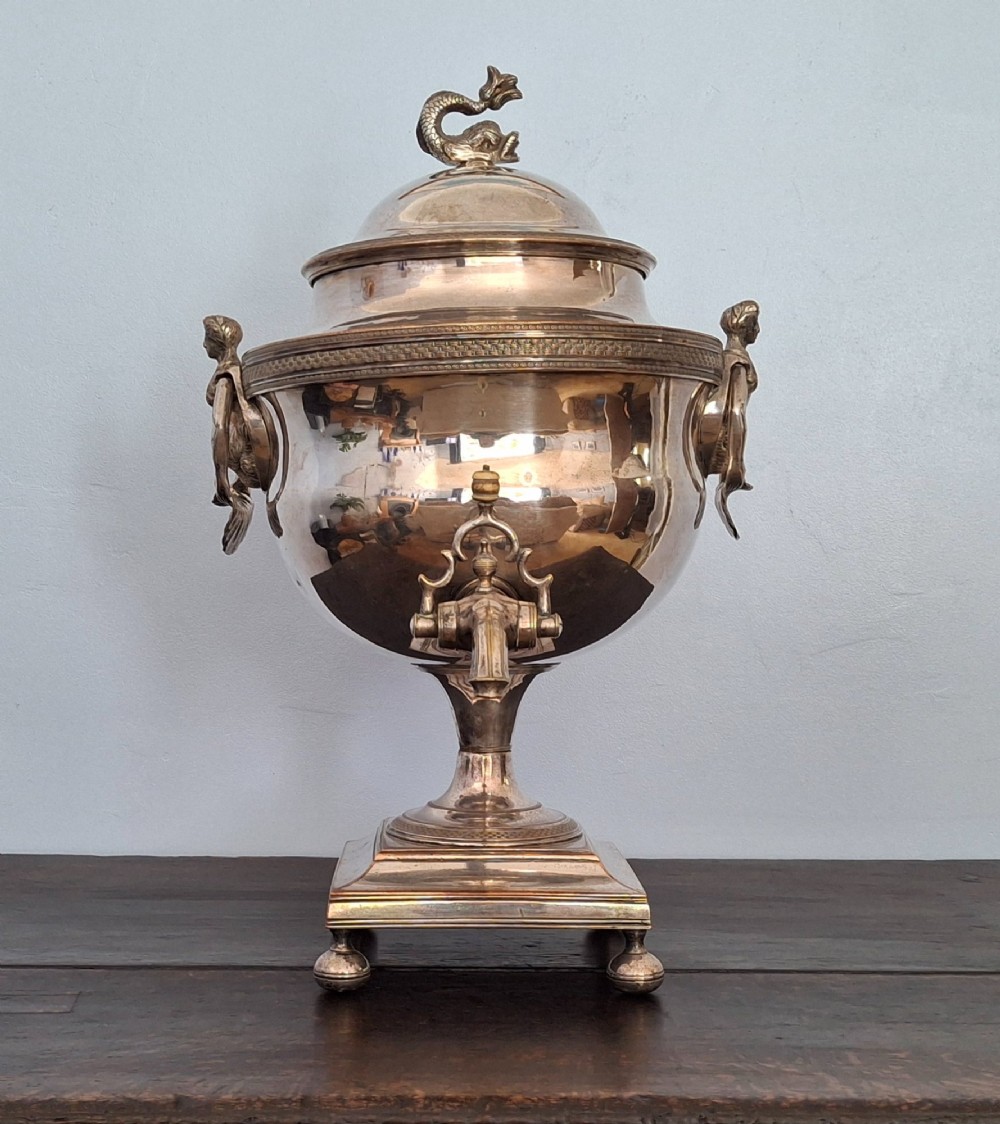 An antique silver-plated Regency tea urn featuring a top dolphin finial, classical figure side handles, a front spigot, and a square base supported by ball feet.