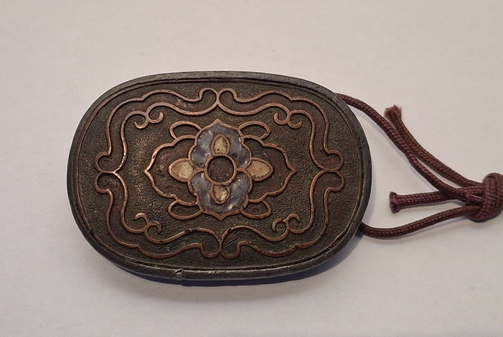 An oval-shaped pewter Japanese manju netsuke featuring champleve enamel floral decoration and an attached cord.