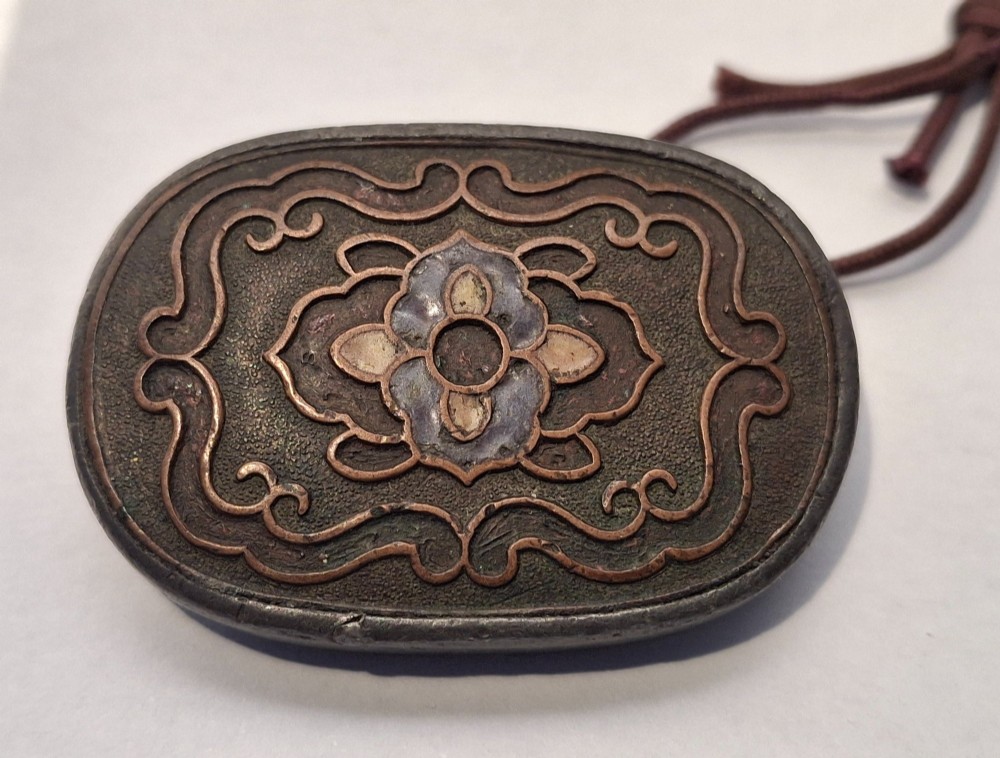 An oval-shaped pewter Japanese manju netsuke featuring champleve enamel floral decoration and an attached cord. View VI