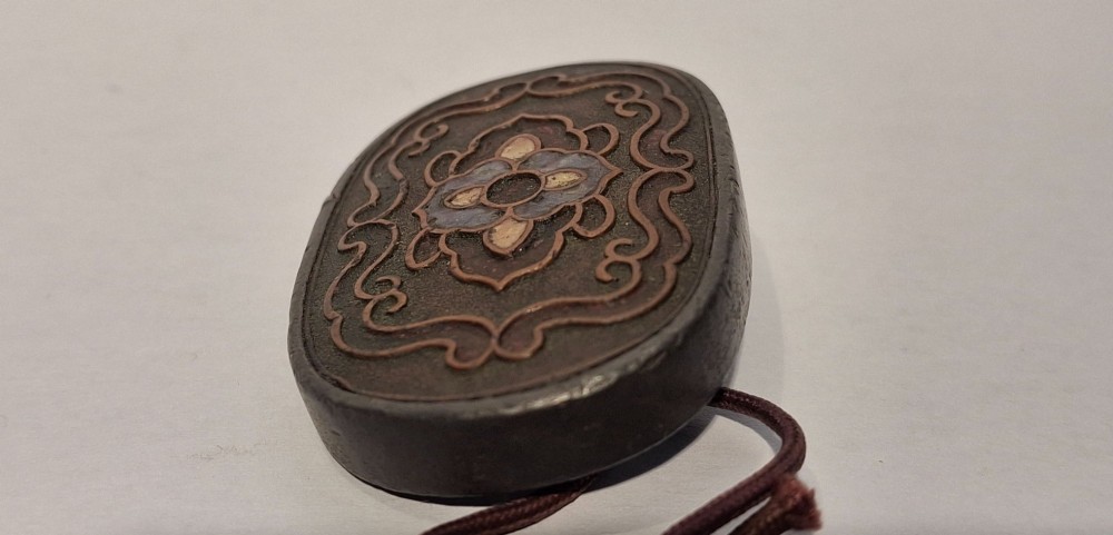 An oval-shaped pewter Japanese manju netsuke featuring champleve enamel floral decoration and an attached cord. View VII