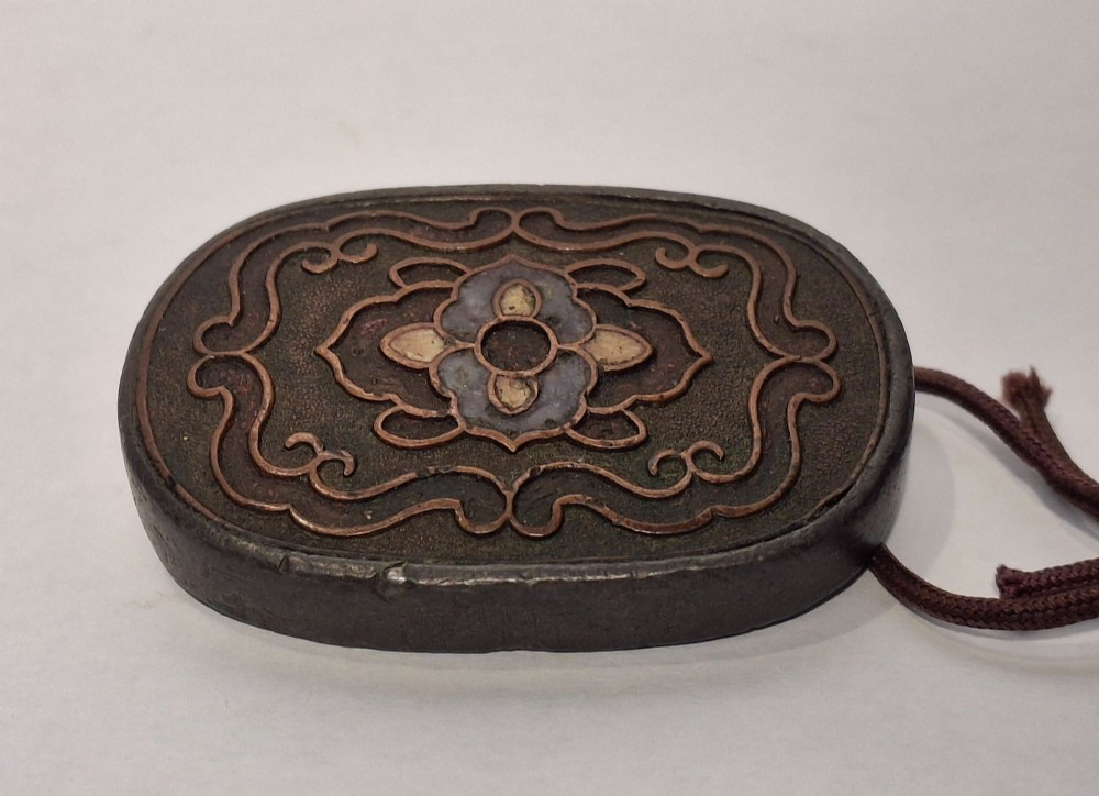 An oval-shaped pewter Japanese manju netsuke featuring champleve enamel floral decoration and an attached cord. View VIII