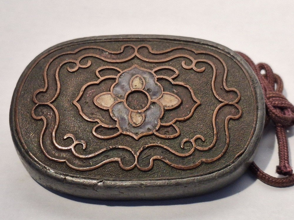 An oval-shaped pewter Japanese manju netsuke featuring champleve enamel floral decoration and an attached cord. View IX