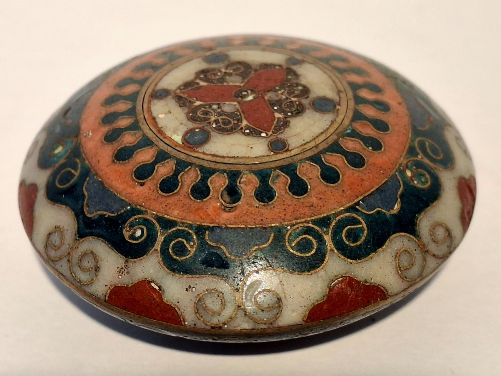 A circular Japanese manju netsuke in polychrome cloisonne enamel with central floral medallion and spiral border patterns.
