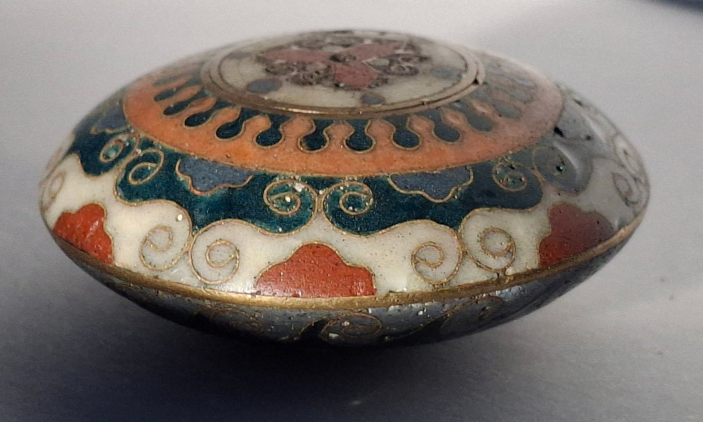 A circular Japanese manju netsuke in polychrome cloisonne enamel with central floral medallion and spiral border patterns. View IX