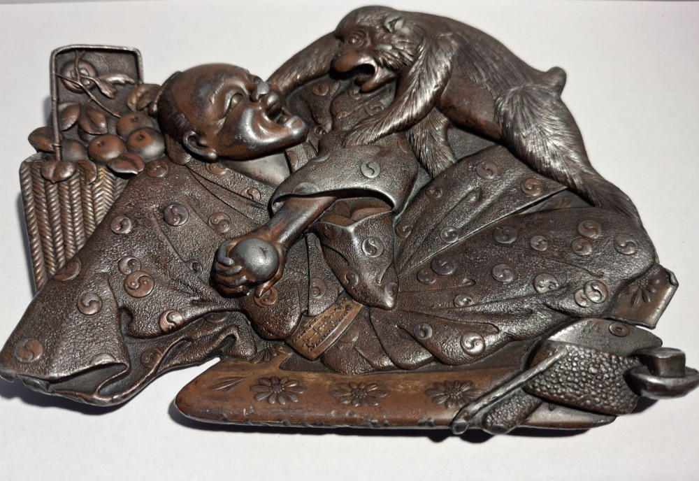 A dark bronze Japanese Takazogan metal tray depicting a figure in patterned robes reacting to a monkey perched on his shoulder.