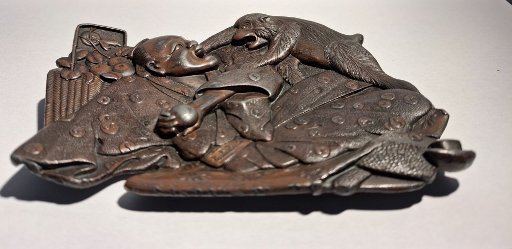 A dark bronze Japanese Takazogan metal tray depicting a figure in patterned robes reacting to a monkey perched on his shoulder. View II