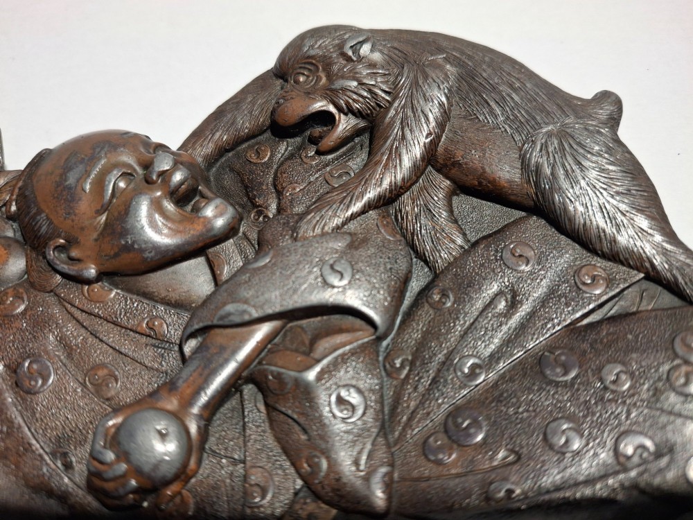 A dark bronze Japanese Takazogan metal tray depicting a figure in patterned robes reacting to a monkey perched on his shoulder. View III
