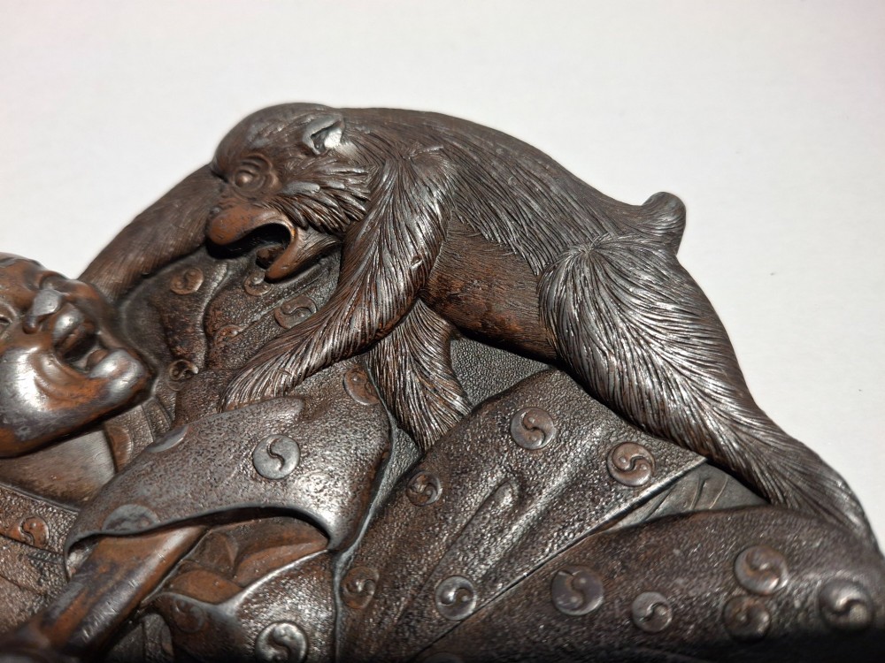 A dark bronze Japanese Takazogan metal tray depicting a figure in patterned robes reacting to a monkey perched on his shoulder. View V