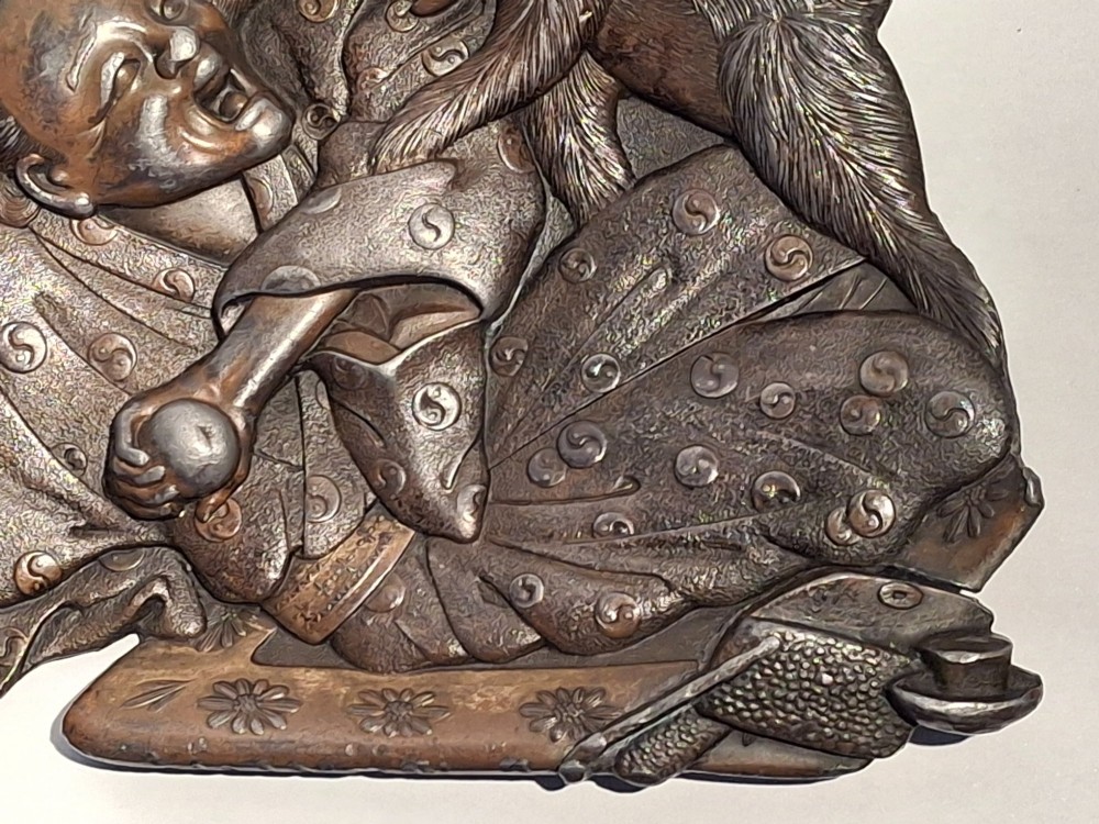 A dark bronze Japanese Takazogan metal tray depicting a figure in patterned robes reacting to a monkey perched on his shoulder. View VI