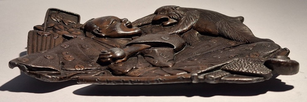 A dark bronze Japanese Takazogan metal tray depicting a figure in patterned robes reacting to a monkey perched on his shoulder. View VIII