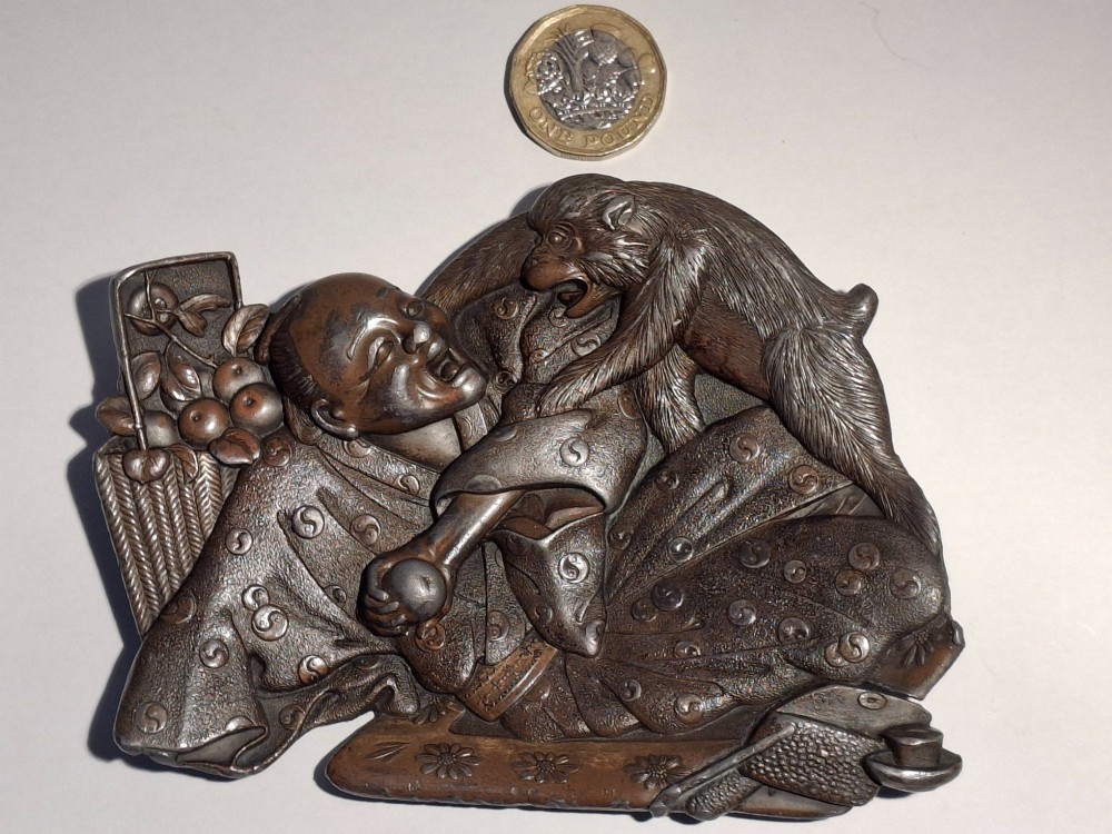 A dark bronze Japanese Takazogan metal tray depicting a figure in patterned robes reacting to a monkey perched on his shoulder. View IX