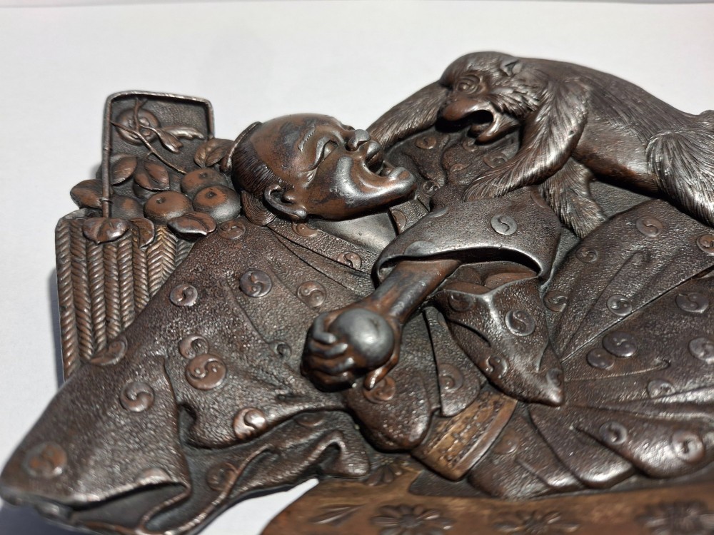 A dark bronze Japanese Takazogan metal tray depicting a figure in patterned robes reacting to a monkey perched on his shoulder. View XII