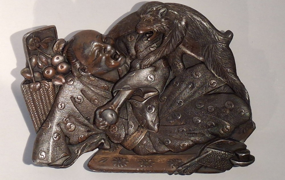 A dark bronze Japanese Takazogan metal tray depicting a figure in patterned robes reacting to a monkey perched on his shoulder. View XIV