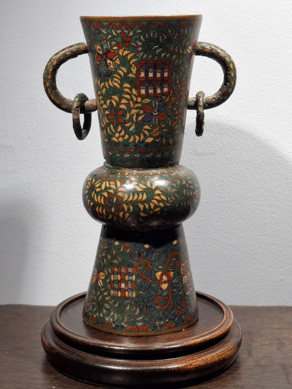 A miniature Japanese cloisonne enamel ritual vase with two loop handles, loose rings, and polychrome floral decoration on a dark green background.