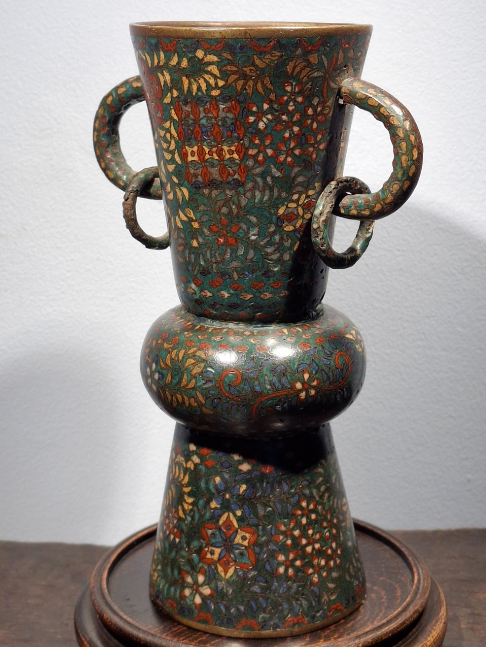 A miniature Japanese cloisonne enamel ritual vase with two loop handles, loose rings, and polychrome floral decoration on a dark green background. View X