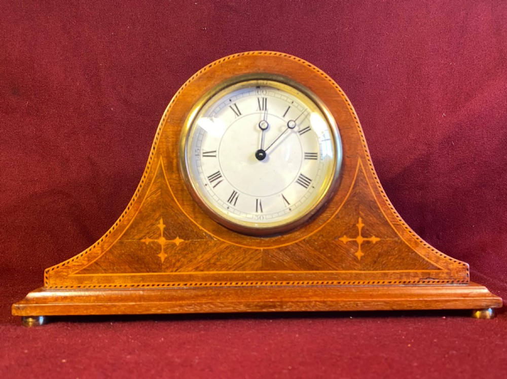 An Edwardian satinwood mantel clock featuring an arched case with geometric cross marquetry, a white Roman numeral dial, and a brass bezel.