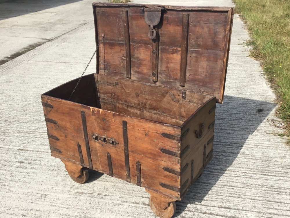 A mid-19th-century colonial wooden strong box featuring wrought iron straps, a central clasp, corner brackets, and large wooden wheels. View V