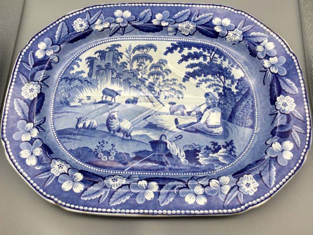 An oval blue and white pearlware platter featuring a printed pastoral scene with sheep, a musician figure, a floral border, and a gravy reservoir well.