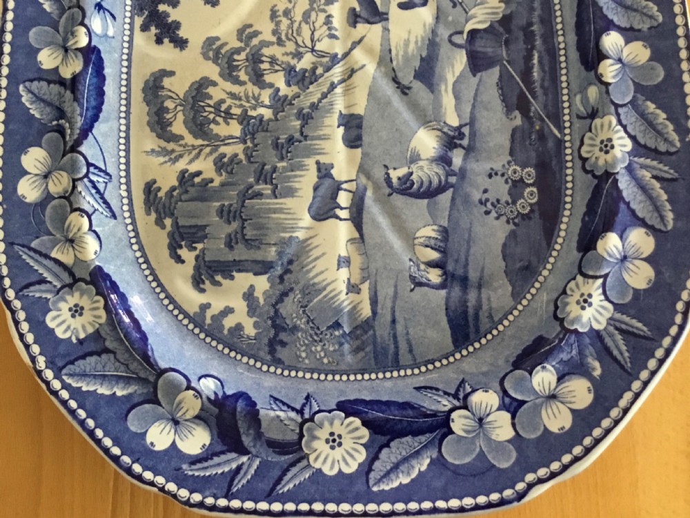An oval blue and white pearlware platter featuring a printed pastoral scene with sheep, a musician figure, a floral border, and a gravy reservoir well. View V