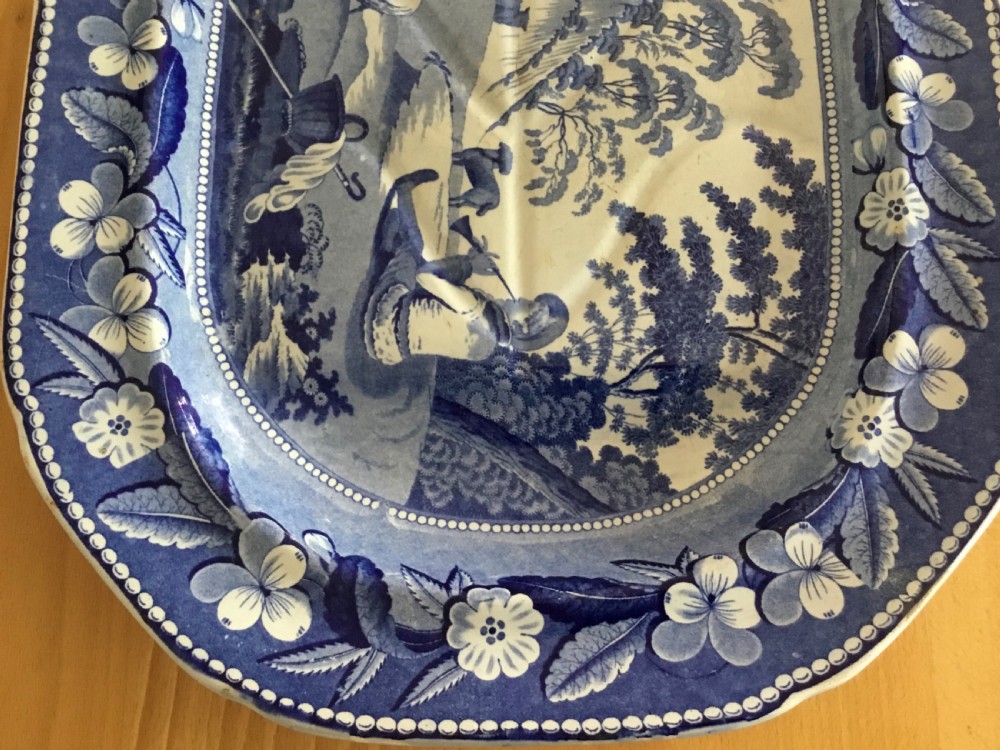 An oval blue and white pearlware platter featuring a printed pastoral scene with sheep, a musician figure, a floral border, and a gravy reservoir well. View VI