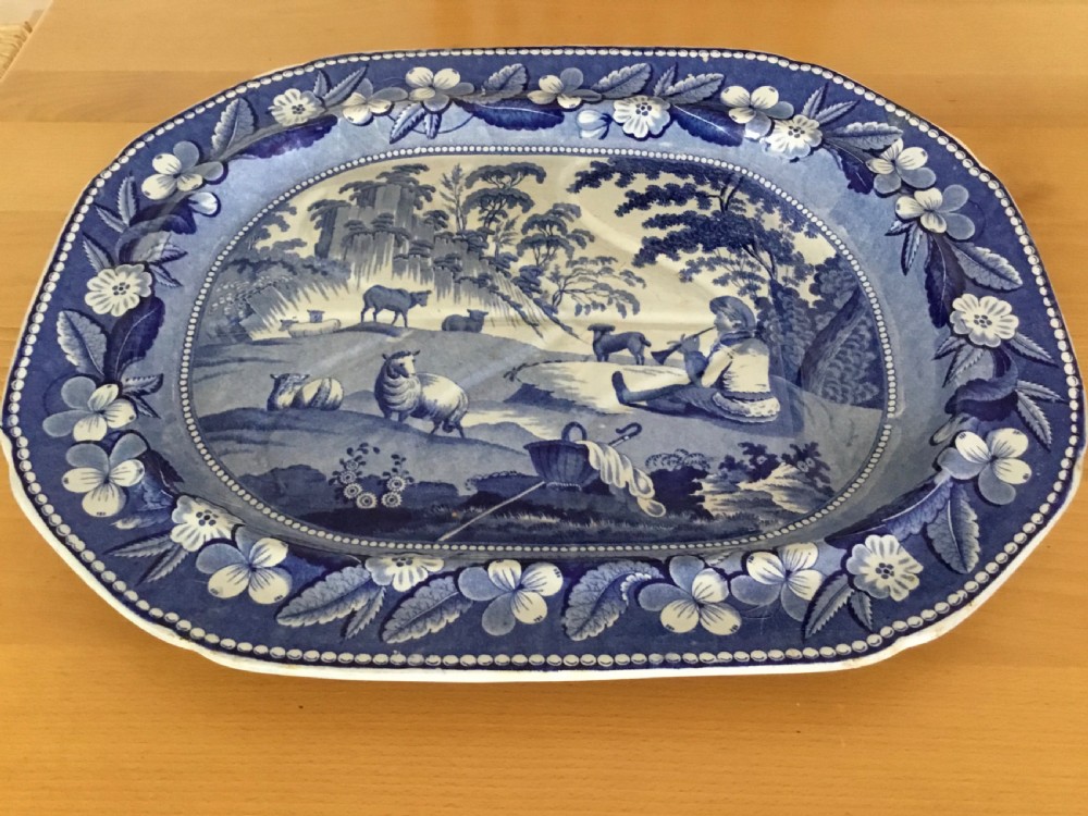 An oval blue and white pearlware platter featuring a printed pastoral scene with sheep, a musician figure, a floral border, and a gravy reservoir well. View VIII