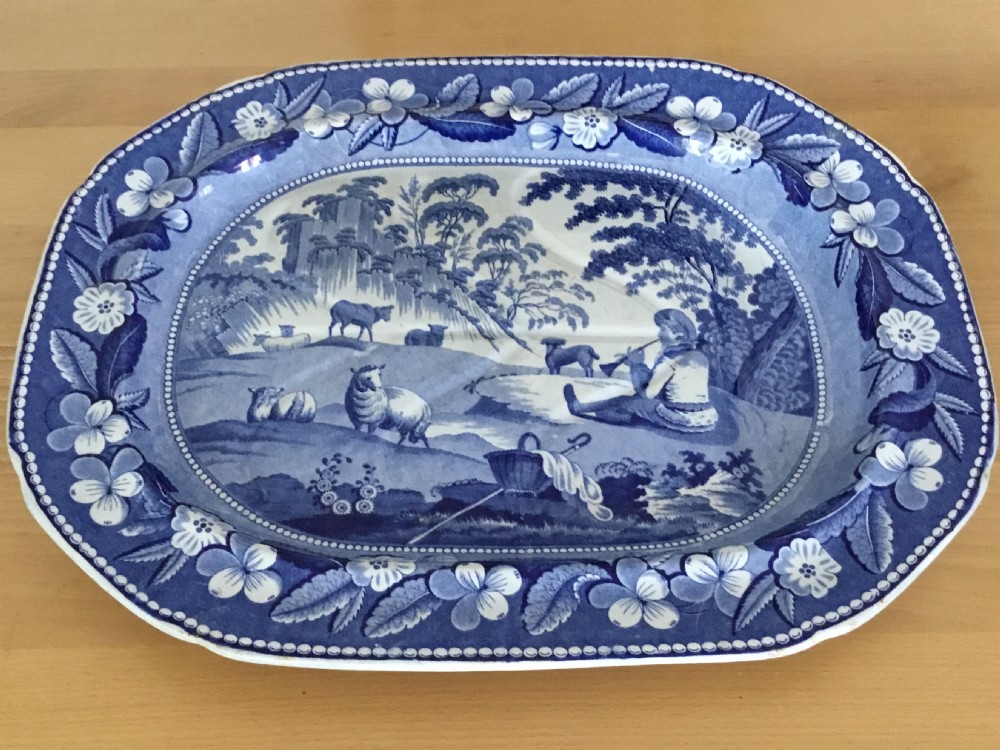 An oval blue and white pearlware platter featuring a printed pastoral scene with sheep, a musician figure, a floral border, and a gravy reservoir well. View IX