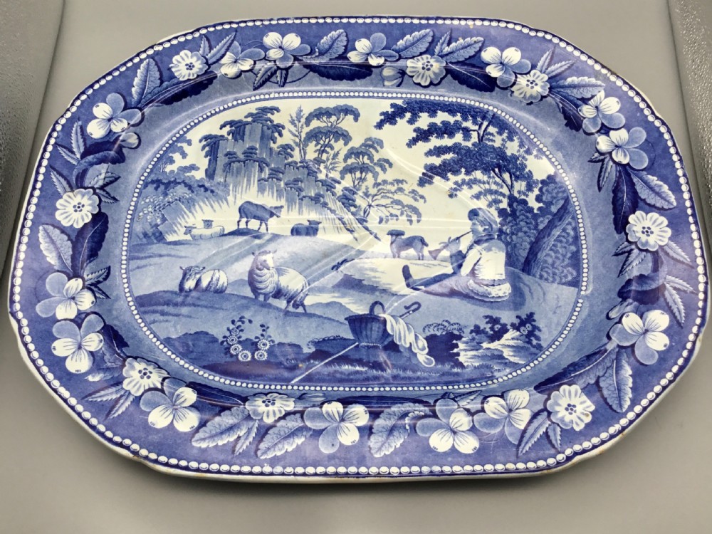An oval blue and white pearlware platter featuring a printed pastoral scene with sheep, a musician figure, a floral border, and a gravy reservoir well. View X