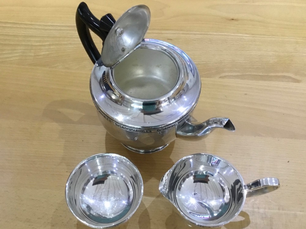 A three-piece silver-plated tea service comprising a teapot with a black curved handle, a sugar bowl, and a milk jug with a band of relief detail along the upper edge. View V